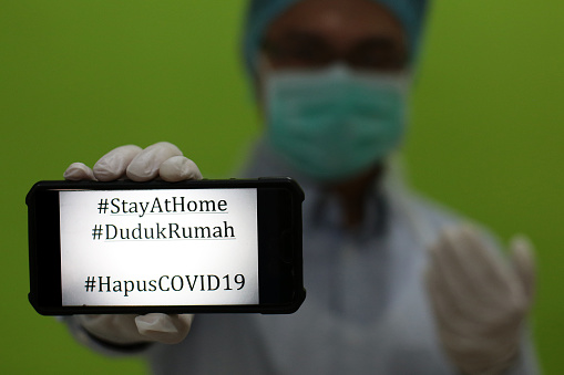 A young man in surgical cap, glove mask and PPE suit is holding a smartphone. It was written with English and Malaysian Covid-19 hashtags messages due to lockdown in Malaysia.