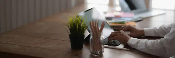 Cropped shot of young college student typing on laptop while sitting in simple workspace with stationery and decoration on wooden desk