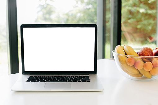 Food blog. Laptop with blank white screen and fruits in glass bowl on countertop   in modern kitchen with big windows, copy space. Culinary courses or recipe online concept. Cooking home concept.