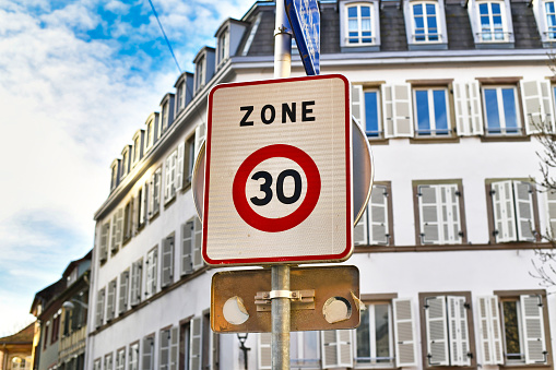 Square shaped white 30km/h speed limit zone sign with red circle in front residential buildings in background in France