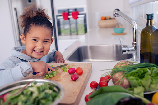 6 years old girl cutting vegetables, preparing a fresh salad. The child looking over the camera with a smile.