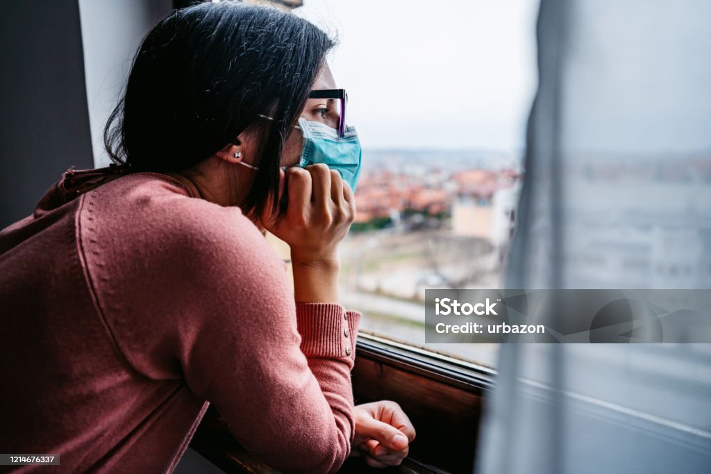 Woman in quarantine looking through the window Woman in quarantine wearing a mask and looking through the window. Pandemic of Coronavirus / Covid-19. Lockdown Stock Photo