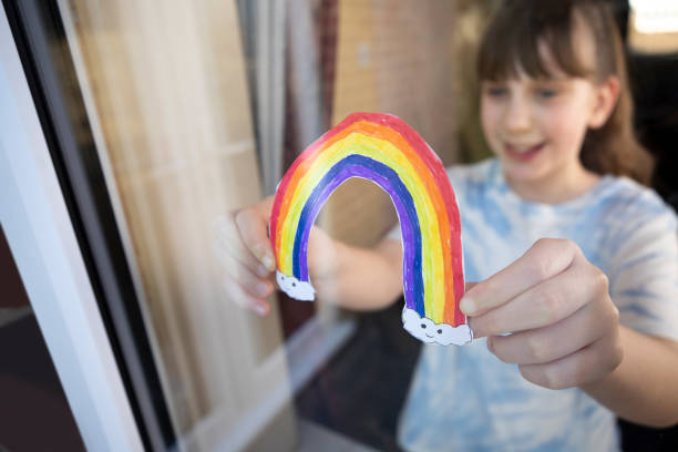 ragazza che mette la foto dell'arcobaleno in finestra a casa durante la pandemia di coronavirus - putting together foto e immagini stock
