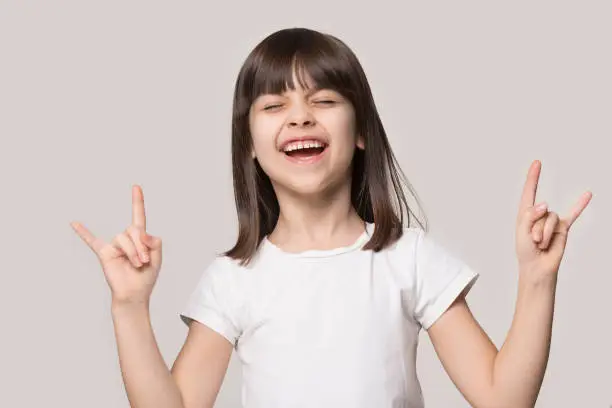 Funny happy small preschooler girl isolated on grey studio background show rock-n-roll gesture relax chill, overjoyed cute little child make death horns sign feel excited uplifted, kid rocker concept