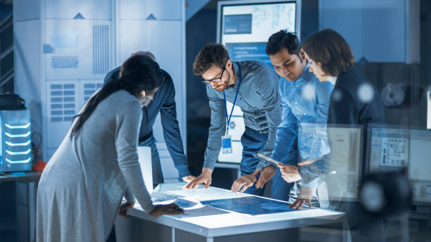istock Engineers Meeting in Technology Research Laboratory: Engineers, Scientists and Developers Gathered Around Illuminated Conference Table, Talking and Finding Solution. Industrial Design Facility 1214717311