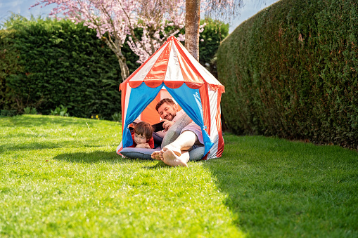 Quarantine and self isolation during coronavirus pandemic period concept. Humour and positive attitude. Man with little son sitting in child tent outdoors in garden at home