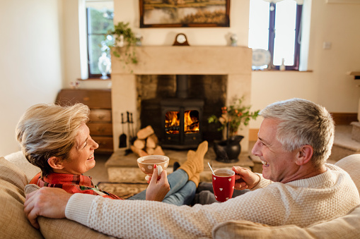 Mature couple sat infront of a fire drinking hot chocalate from mugs.