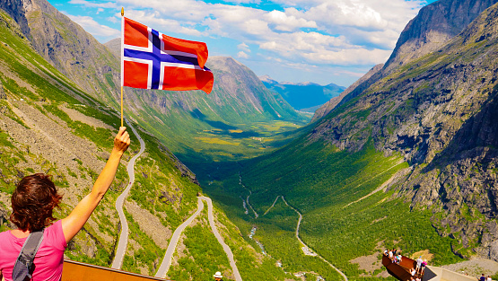 Tourist woman with norwegian flag on Trollstigen viewpoint. People on viewing platform in the background. Trolls Path mountain road in Norway. National route. Touristic attraction.