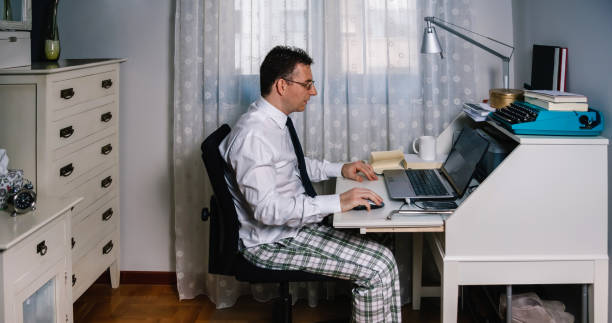 Man teleworking wearing shirt, tie and pajama pants Man working from home with laptop wearing shirt, tie and pajama pants desk lamp photos stock pictures, royalty-free photos & images