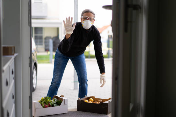 neighborly help during covid-19, friendly waving woman delivering fresh food friendly waving helpful mature adult woman with protective gloves, goggles and mask at open door of private house delivering fresh food in box during covid-19 coronavirus crisis curfew quarantine, shallow focus curfew stock pictures, royalty-free photos & images