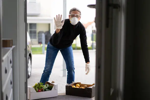 friendly waving helpful mature adult woman with protective gloves, goggles and mask at open door of private house delivering fresh food in box during covid-19 coronavirus crisis curfew quarantine, shallow focus