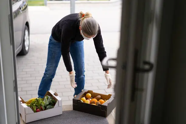 mature adult woman with protective gloves and goggles and respiratory mask at open door of private house delivering fresh food in box during covid-19 coronavirus crisis curfew quarantine, shallow focus