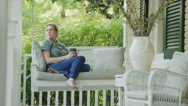 A Caucasian Woman in her Forties Drinks a Cup of Coffee while Sitting on a Porch Swing Outdoors Surrounded by Lush Greenery
