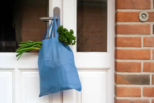 Blue shopping bag with fresh vegetables and goods was hanged on the front door, help concept during quarantine time because of coronavirus infection, copy space, selected focus