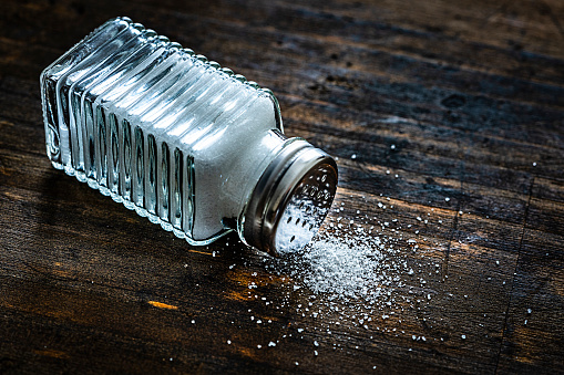 Close up view of a glass salt shaker shot on rustic wooden table. The shaker is laying down and some salt is spilled on the table. The composition is at the left of an horizontal frame leaving useful copy space for text and/or logo at the right. Predominant colors are white and brown. High resolution 42Mp studio digital capture taken with Sony A7rII and Sony FE 90mm f2.8 macro G OSS lens
