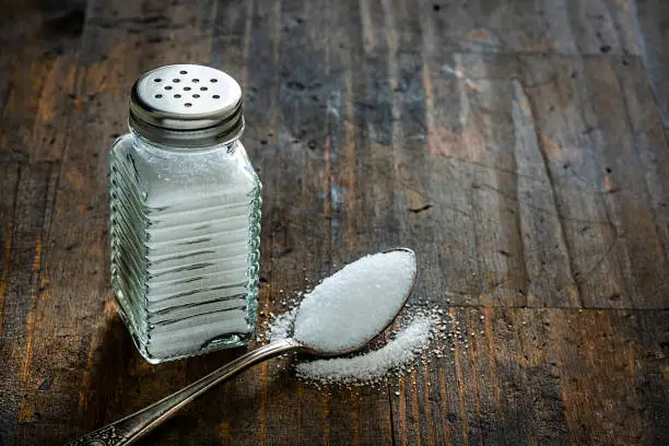 Glass salt shaker shot on rustic wooden table. A spoon with salt is beside the shaker placed directly on the table. The composition is at the left of an horizontal frame leaving useful copy space for text and/or logo at the right. Predominant colors are white and brown. High resolution 42Mp studio digital capture taken with Sony A7rII and Sony FE 90mm f2.8 macro G OSS lens