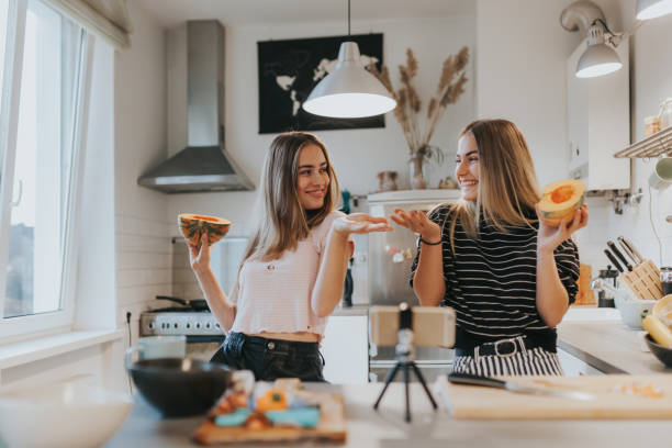 Two teenage girlfriends filming a music video in the kitchen with smart phone Photo series of two female teenage vegan vloggers filming a show about cooking vegan foods. cooking show stock pictures, royalty-free photos & images