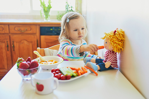 Adorable toddler girl eating fresh fruits and vegetables for lunch. Child feeding doll and teddy bear in the kitchen. Delicious healthy food for kids