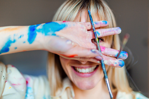 Portrait of a beautiful young blonde woman covered in colors, covering her face and holding a paintbrush