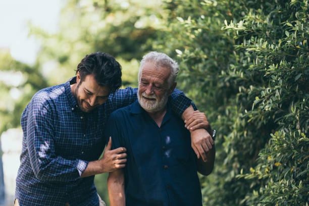 de oude man en zijn zoon lopen in het park. een man knuffelt zijn bejaarde vader. ze zijn blij en glimlachend - zonlicht fotos stockfoto's en -beelden