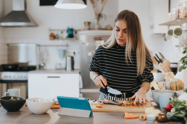 Cute female teenager cooks dinner while having a video call conversation Photo series of two female teenage vegans learning to cook while watching tutorials in internet. cooking stock pictures, royalty-free photos & images