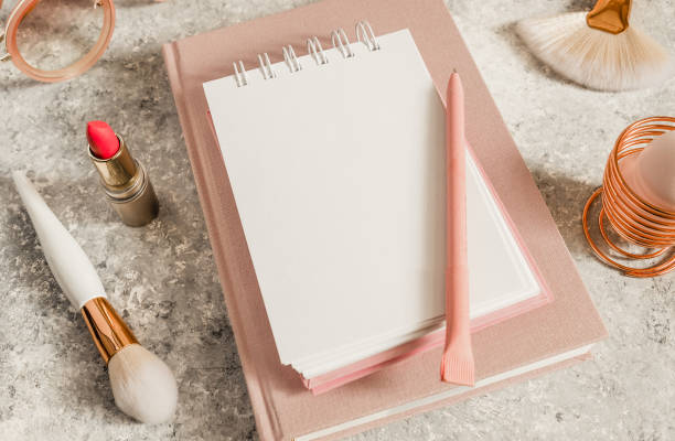 istock Female workspace with paper notebook, pink flowers, coffee cup, lipstick, makeup brushes and sunglasses. Selective focus. Natural soft light from the window 1214769131