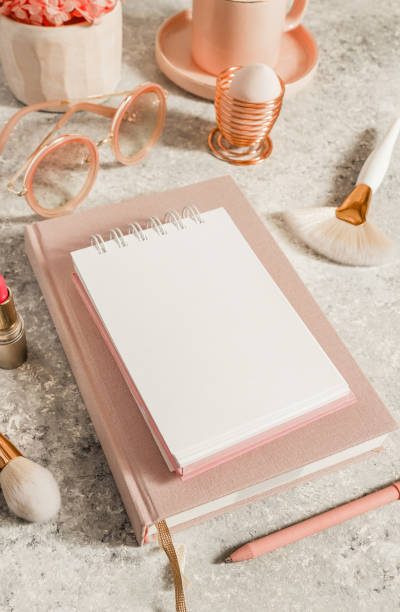 istock Female workspace with paper notebook, pink flowers, coffee cup, lipstick, makeup brushes and sunglasses. Selective focus. Natural soft light from the window 1214769148