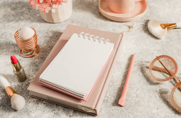 istock Female workspace with paper notebook, pink flowers, coffee cup, lipstick, makeup brushes and sunglasses. Selective focus. Natural soft light from the window 1214769164