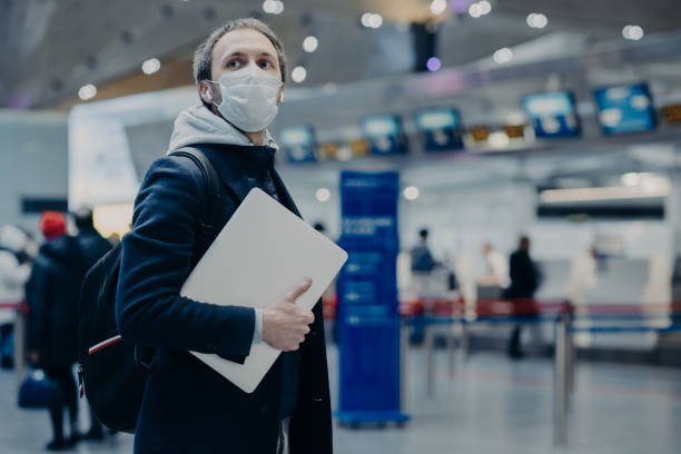 Man traveler wears protective disposable medical mask in airport, returns from abroad where coronavirus spreading, carries backpack, takes care of health, protects from virus, has postponed flight Man traveler wears protective disposable medical mask in airport, returns from abroad where coronavirus spreading, carries backpack, takes care of health, protects from virus, has postponed flight human cardiopulmonary system audio stock pictures, royalty-free photos & images