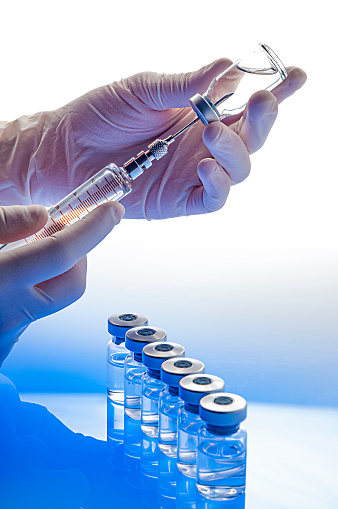 Preparing vaccine: Hands with gloves holding syringe and vial. Blue toned image. Selective focus. Several vials are out of focus on background on foreground and two vials are out of focus at background