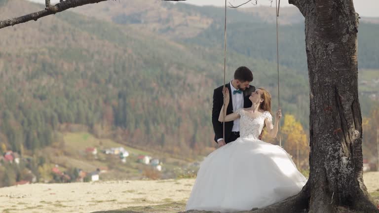 Newlyweds. Caucasian groom with bride ride a rope swing on a mountain slope