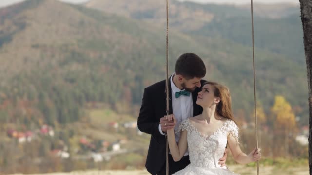 Newlyweds. Caucasian groom with bride ride a rope swing on a mountain slope