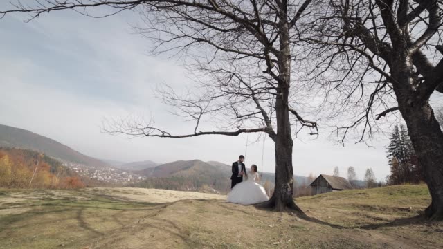 Newlyweds. Caucasian groom with bride ride a rope swing on a mountain slope