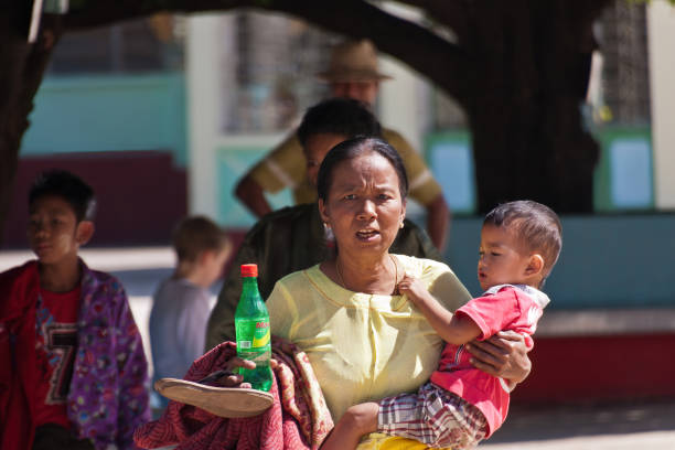 A Burmese woman is hurrying somewhere holding a bottle with a drink in one hand and a child in another.