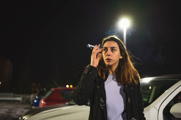 Beaten young woman, smoking cigarette at the parking lot near car to relax herself after she escape her abuser
