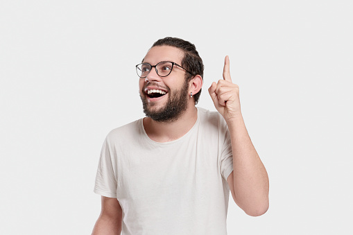 Portrait of a excited young man with glasses pointing up with cheerful facial expression over graybackground. Studio shot. Horizontal composition.