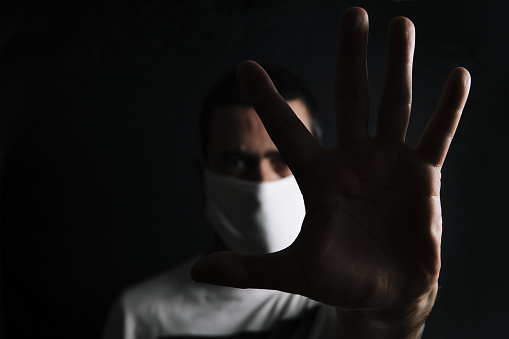 dark portrait of a man with a protective mask doing stop sign with the hand in black background, selective focus, concept of coronavirus and protection against viruses and bacteria