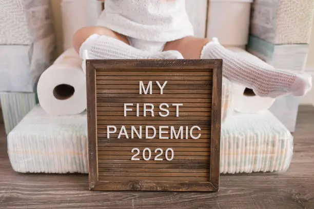 A comical depiction of a baby's first pandemic during the Covid-19 virus. Letterboard sign saying "My First Pandemic 2020" with quarantine essentials in the background like towel paper, tissues, diapers, wipes & paper towels.