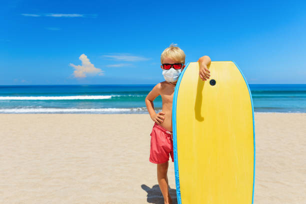 Young surfer wearing sunglasses, protective mask on sea beach Young surfer wearing sunglasses, protective mask on sea beach with body board. Summer tours, cruises cancellation due to coronavirus COVID-19 epidemic. Safe travel destinations for family vacation. body board stock pictures, royalty-free photos & images