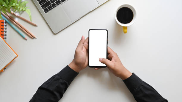 istock Top view image of hands holding a cropped black smartphone with white blank screen over a coffee cup, computer laptop, pencil, notebook putting on working desk as background. 1214852674