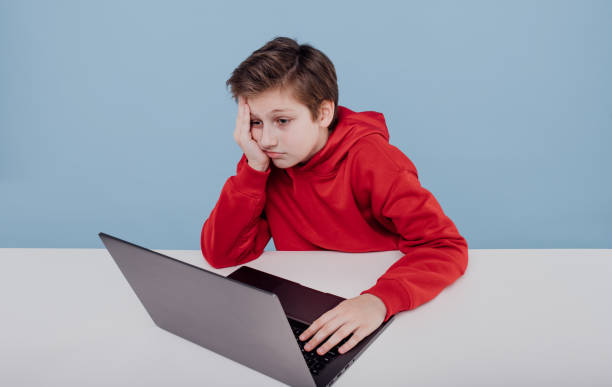 istock tired boy in red sweatshirt near laptop in studio on blue background 1214857323