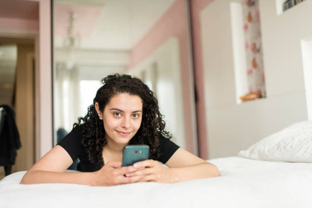 adolescente sorridente sdraiata sul suo letto e guardando la telecamera - one teenage girl only on the phone bed lying on front foto e immagini stock