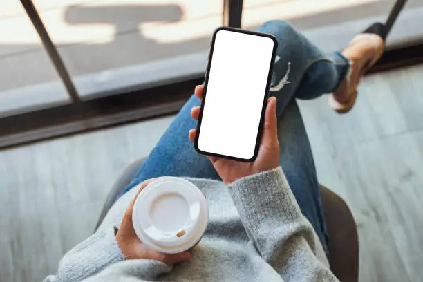 Photo of a woman holding a black mobile phone with blank white desktop screen with coffee cup