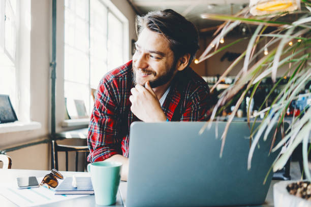 istock Young stubbled man working with laptop in big bright office space. Smiling and thinking about new idea 1214876046
