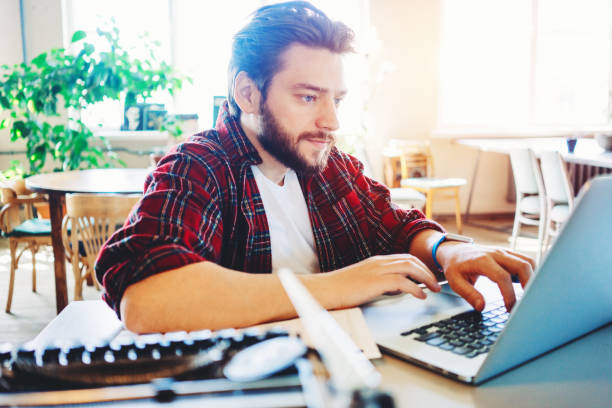 istock Man with stubble and flannel shirt working with laptop in bright office space 1214877593