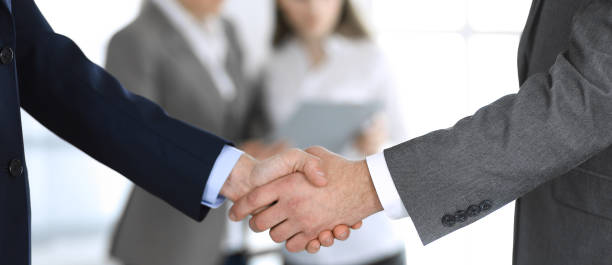 istock Business people shaking hands while standing with colleagues after meeting or negotiation, close-up. Group of unknown businessmen and women in modern office. Teamwork, partnership and handshake concept, toned picture 1214886484