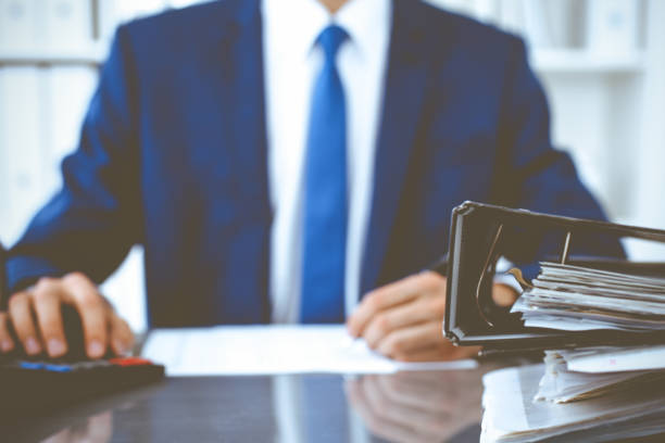 istock Binders with papers waiting to be processed with businessman or bookkeeper back in blur. Accounting planning budget. Audit, insurance and business concept 1214886670