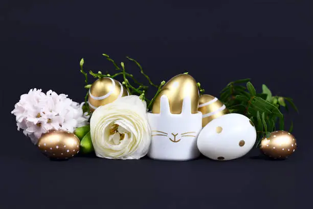 Modern metallic easter eggs painted with metallic golden and white stripes and dots and cute easter egg cup in shape of bunny surrounded by spring flowers like cream colored buttercup flower on dark black background