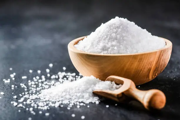 Photo of Salt. Glass salt shaker on dark stone table.  A pile of salt top view and wooden bowl