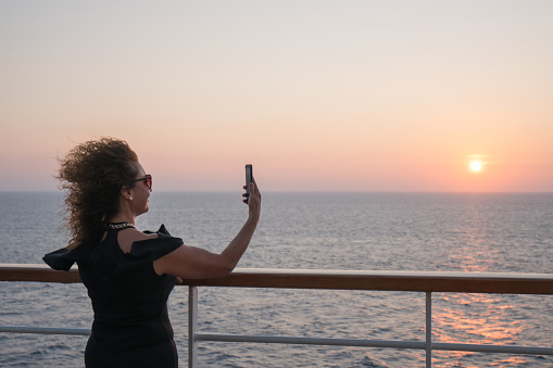 Beautiful woman standing on deck of cruise ship and doing selfie at sunset.